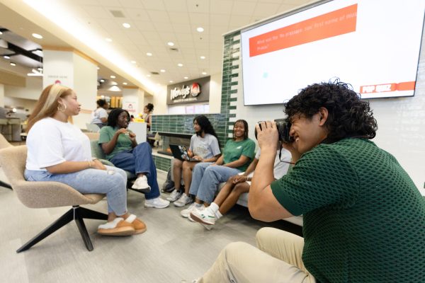 smiling student intern wearing a green polo holds a camera to his eye, taking a picture of a group of students sitting in a circle in the Student Union food court
