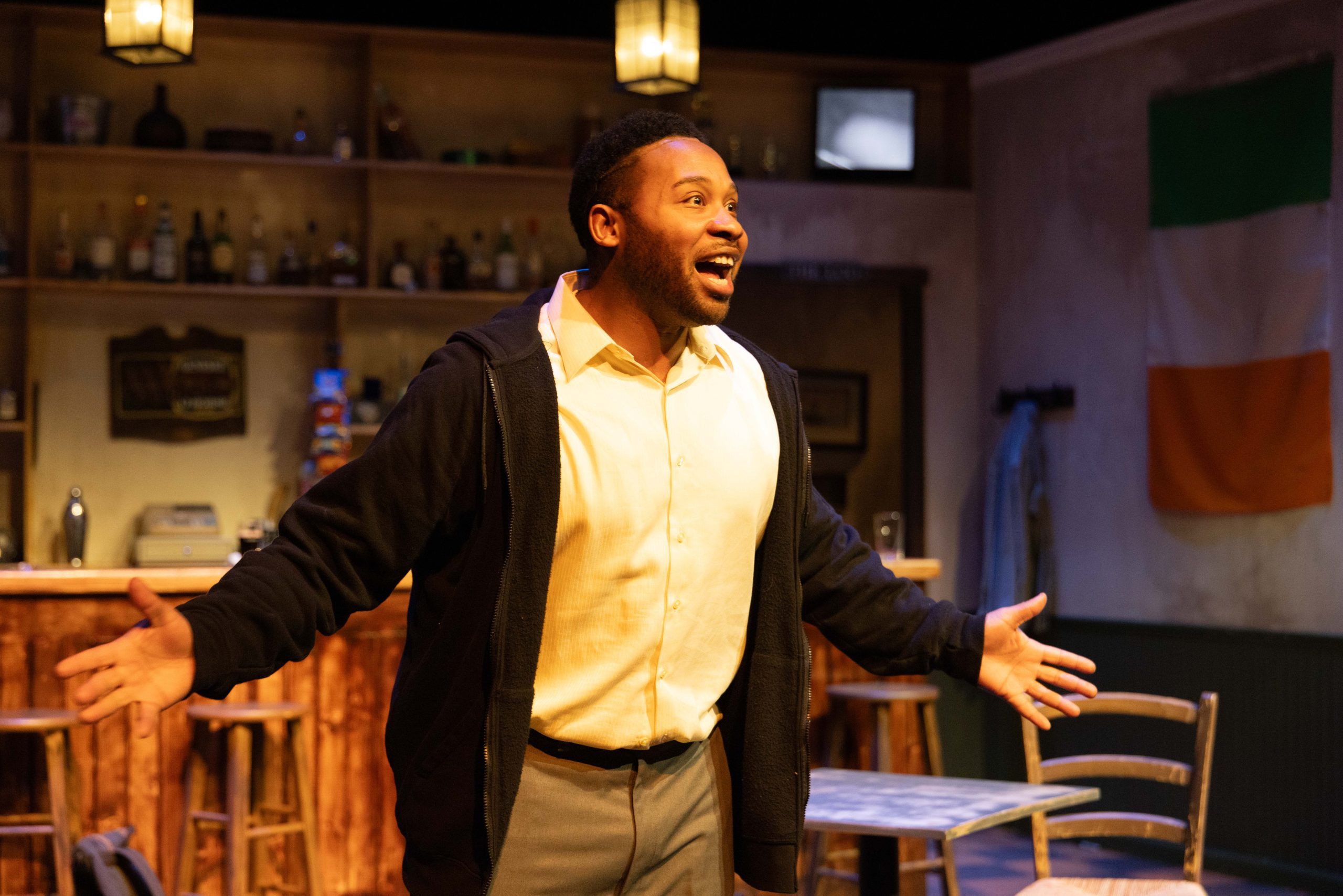 a male student on stage in a play with his arms wide open talking to the audience 